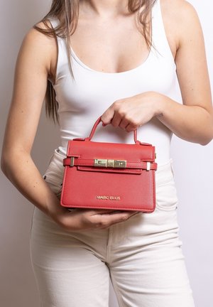 Femme en débardeur blanc et pantalon tenant un sac à main rouge Marc Ellis avec des ferrures dorées au niveau de la taille.