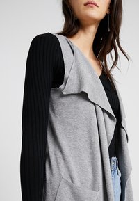 Cardigan grigio con apertura frontale, realizzato in un tessuto morbido, caratterizzato da reverse drappeggiati e due tasche frontali, abbinato a una maglietta nera a maniche lunghe a costine.