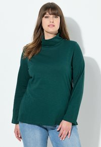 Maglia a maniche lunghe color verde scuro con collo alto realizzata in tessuto morbido, caratterizzata da una vestibilità comoda e un design semplice, abbinata a jeans in denim blu chiaro.