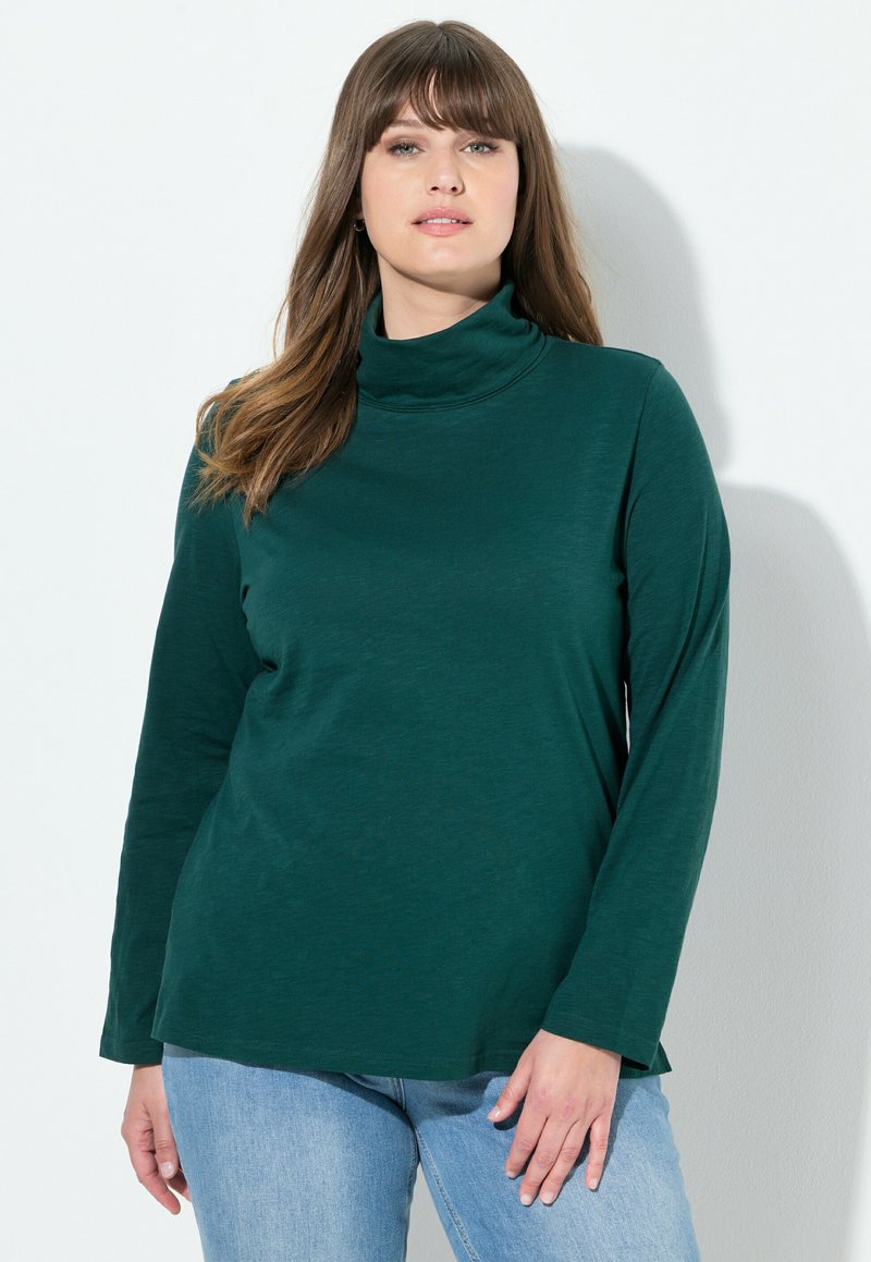 Maglia a maniche lunghe color verde scuro con collo alto realizzata in tessuto morbido, caratterizzata da una vestibilità comoda e un design semplice, abbinata a jeans in denim blu chiaro.