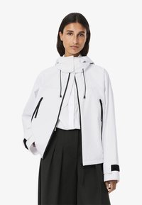 Giacca impermeabile bianca con colletto alto, lacci regolabili e tasche con zip nere. Presenta una vestibilità rilassata e oversize.