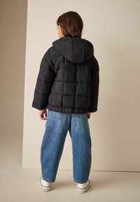 Czarna pikowana kurtka puchowa z kapturem o pudełkowym kroju. Nosi się ją na niebieskich spodniach o szerokich nogawkach i białych trampkach.