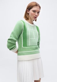 Jeune femme portant un pull texturé vert et blanc à motifs, associé à une jupe plissée blanche, debout devant un fond blanc uni.