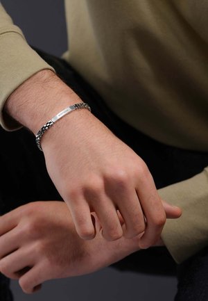 Zilveren metalen armband met zwarte kettingschakels, met bovenop een platte, rechthoekige graveerschijf. De textuur is glad en glanzend.