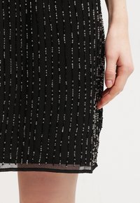 Czarna sukienka z pionowymi koralikowymi akcentami. Przezroczysta tekstura z warstwowym wykończeniem u dołu. Ręka umiejscowiona blisko dolnej krawędzi.