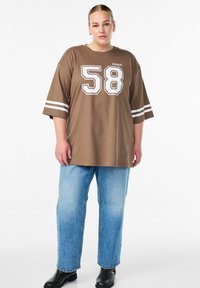 Rjava prevelika majica s kratkimi rokavi z belim potiskom "58" in črtami na rokavih. Kombinirana z svetlo modrimi kavbojkami z ravnimi nogami in črnimi gležnarji.