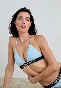 Světle modrý trojúhelníkový bikiny top s tenkými ramínky, obsahující černý značkový pásek. Hladká textura, minimalistický design a kontrastní barvy.