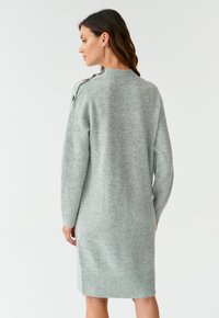 Robe pull tricotée gris clair, avec un ourlet et des poignets côtelés, dotée d'un détail de bouton sur l'épaule, tombe à mi-cuisse.