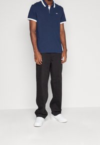 Námořnická polo košile s bílým límcem a akcenty na rukávech, v kombinaci s černými kalhotami a bílými teniskami, ukazující volný střih a jednoduchý design.
