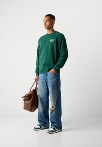 Groene sweatshirt, losse lichtblauwe jeans met een rafelrand en groen/witte sneakers. Bruine leren tas met bovenhandvatten. Eenvoudig ontwerp.