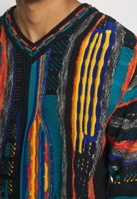 Neulepaita, jossa on eläväinen kuviointi, jossa on raitoja mustassa, punaisessa, keltasessa, sinisessä ja turkoosissa. V-kaula-aukko ja tekstuuriset yksityiskohdat.