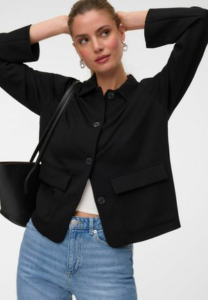 Femme portant une veste noire boutonnée sur un top blanc avec un jean bleu, portant un sac à épaule noir, posant avec les mains derrière la tête.