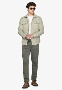 Lys grå uld skjortejakke med to brystlommer, båret over en hvid skjorte. Olivengrønne bukser og brune snørresko fuldender outfittet.