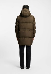 Puffermantel in dunkel olivgrün, mit Kapuze, gestepptem Muster und elongated Silhouette. Kombiniert mit schwarzen Hosen und Schuhen.
