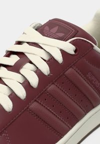 Μπορντό αθλητικό παπούτσι με λείο δερμάτινο επάνω μέρος, λευκά κορδόνια, τρεις λωρίδες στο πλάι, μικρές τρύπες για αερισμό και ανάγλυφο λογότυπο στη γλώσσα.
