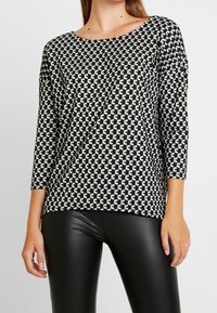 Langarmoberteil mit schwarz-weißer Musterung und Rundhalsausschnitt, das über ein geometrisches Design und eine lässige Passform verfügt, kombiniert mit schwarzen Leggings.