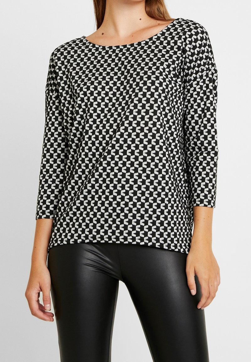 Langarmoberteil mit schwarz-weißer Musterung und Rundhalsausschnitt, das über ein geometrisches Design und eine lässige Passform verfügt, kombiniert mit schwarzen Leggings.