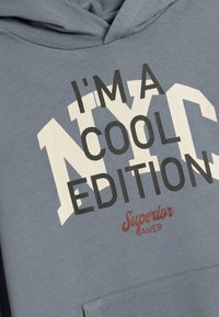 Felpa grigia con cappuccio con grande testo grafico bianco e nero che dice "SONO UNA VERSIONE COOL DI NYC" con "Superior Gamer" in rosso. Tasca a canguro frontale.