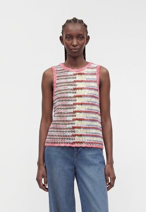Femme aux cheveux tressés portant un haut crochet multicolore sans manches avec bordure rose et jean bleu large sur un fond uni.
