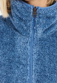 Närbild av en blå fluffig fleecetröja med framzipper och hög krage som ligger över personens hals och haka.
