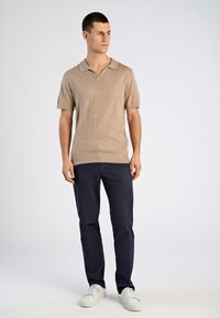 Beige kortärmad polo med krage, texturerad tyg, kombinerad med marinblå raka byxor och vita sneakers, stående framför en ljus bakgrund.