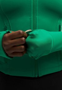 Gros plan sur des mains à la peau foncée ajustant le poignet à trou pour le pouce d'une veste à fermeture éclair vert vif avec un pantalon noir.