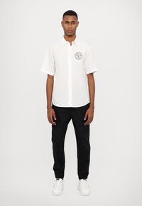 Chemise blanche à manches courtes à boutons avec un logo circulaire noir sur la poitrine gauche, associée à un pantalon noir et des baskets blanches.