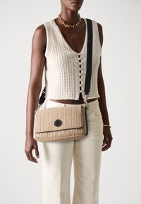 Woven beżowa torba na ramię z zapięciem na klapkę i czarnymi akcentami. Posiada okrągłe logo oraz regulowany pasek, w komplecie z teksturowaną dzianinową bluzką.