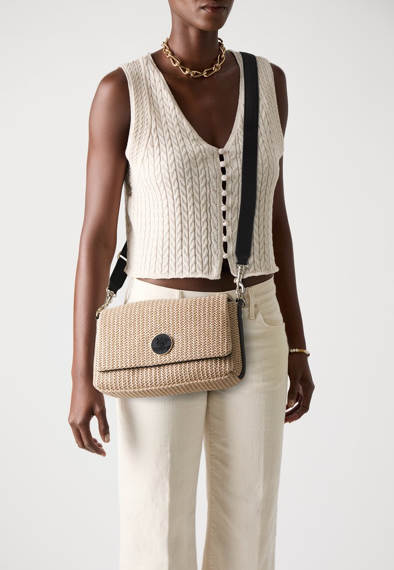 Woven beżowa torba na ramię z zapięciem na klapkę i czarnymi akcentami. Posiada okrągłe logo oraz regulowany pasek, w komplecie z teksturowaną dzianinową bluzką.