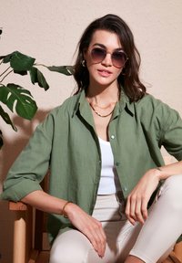 Green linen shirt with rolled sleeves, worn over a white tank top. Model wears round sunglasses and gold jewelry. Light beige pants.