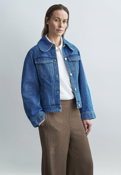 Giacca di denim con ampio colletto, tasche frontali e chiusure con bottoni in blu medio. Indossata sopra una camicia bianca e pantaloni marroni con trama.