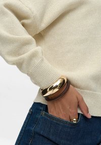 Hellbeige gestrickter Pullover mit gerippten Bündchen, kombiniert mit einem braunen Leder- und goldfarbenen Metallarmband am Handgelenk, Hände in der Denim-Tasche.