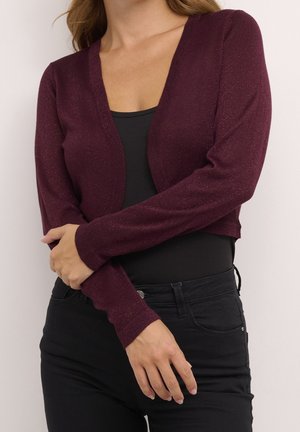 Femme portant un cardigan court à manches longues de couleur violet foncé par-dessus un haut noir et un pantalon noir, debout les bras croisés devant elle.
