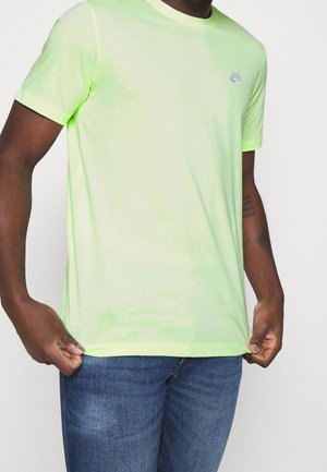 Mann trägt ein hellgrünes Nike-T-Shirt und blaue Jeans, hält den Saum des T-Shirts mit beiden Händen.