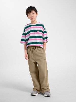 Junge steht mit Händen in den Taschen und trägt ein grün, rosa und weiß gestreiftes Hemd, beige Hose und graue Sneakers vor weißem Hintergrund.