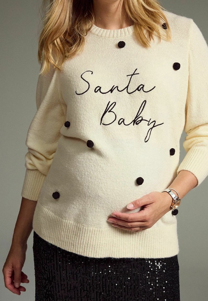 Pull crème avec pompons noirs, arborant le texte "Santa Baby" en cursive. Porté avec une jupe noire à sequins, silhouette ajustée.