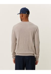 Maglione lavorato a maglia beige chiaro con maniche lunghe, scollatura rotonda e polsi a coste, indossato con pantaloni scuri e un berretto blu navy.
