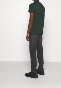 Hombre con camiseta verde oscura, jeans gris oscuro y zapatillas negras, de pie de lado frente a un fondo blanco liso.