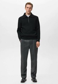 Maglione nero lavorato a maglia con colletto a mezza zip, abbinato a pantaloni grigi formali e scarpe nere. Lo sfondo neutro mette in evidenza i dettagli dell'outfit.