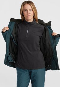 Black fleece pullover with a quarter-zip collar, worn beneath a teal outer jacket; features zippered pockets and a smooth texture.