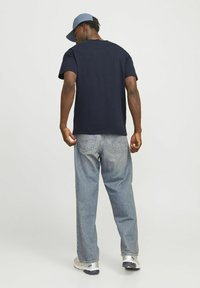 Marineblauwe katoenen T-shirt met korte mouwen, gecombineerd met lichtblauwe baggy jeans. Het model draagt een lichtblauwe pet en heeft een ontspannen houding.