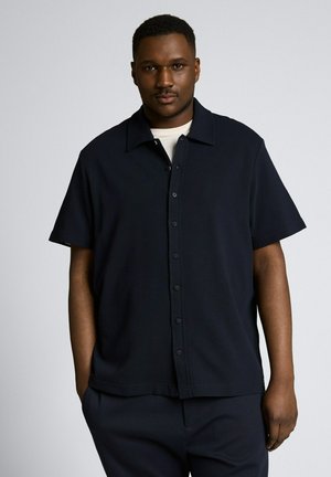 Man met kort haar die een zwart kort mouwen overhemd met knopen draagt over een wit T-shirt, staand tegen een effen lichte achtergrond.