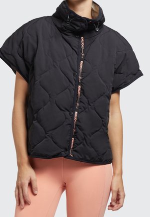 Person iført en kortærmet sort quiltet jakke med hætte og ferskenfarvede leggings mod en ensfarvet baggrund.
