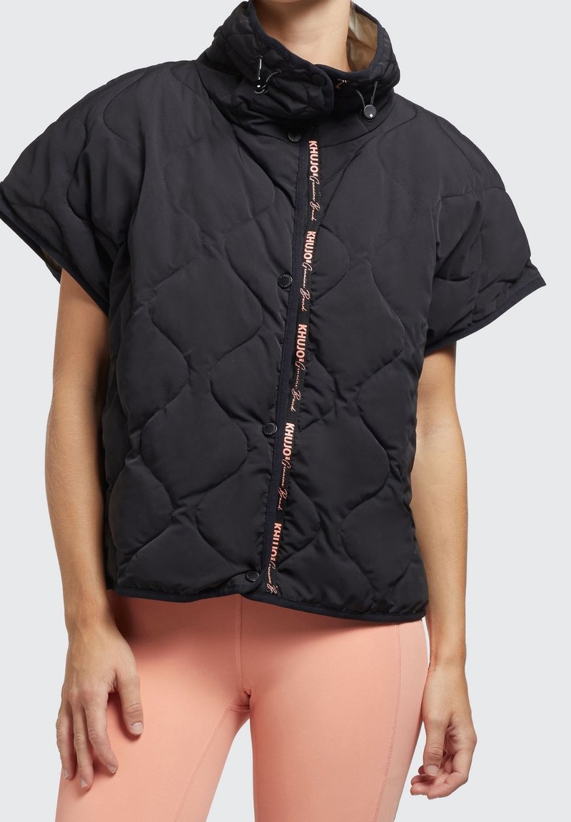 Personne portant une veste matelassée noire à manches courtes avec une capuche et des leggings pêche sur un fond uni.