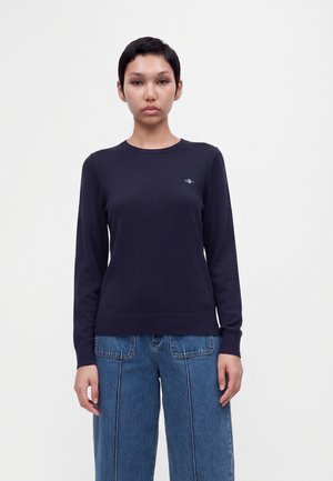 Vrouw met kort donker haar die een aansluitend marineblauw shirt met lange mouwen en blauwe wijde broekspijpen draagt, staat tegen een effen witte achtergrond.