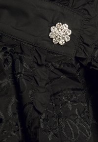 Robe en tissu noir avec des motifs floraux brodés, ornée d'une broche en forme de fleur sertie de bijoux. La texture plissée met en valeur le design.