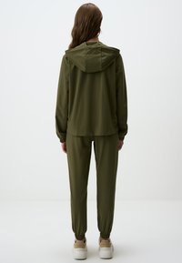 Survêtement de sport vert olive composé d'une veste légère et d'un pantalon assorti. La veste est dotée d'une capuche ; le pantalon a des poignets élastiques. Design décontracté.