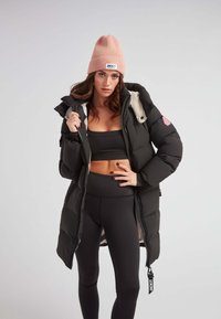 Chaqueta acolchada negra con forro blanco, que presenta un gorro de canalé rosa. La chaqueta tiene una cremallera, bolsillos laterales y una etiqueta de logo. Leggings negros completan el conjunto.