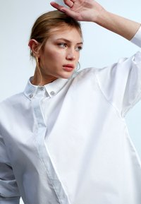 Camicia bianca con colletto a punta, caratterizzata da decorazioni floreali in argento sul colletto e una texture morbida e leggera.