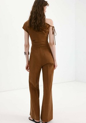 Femme aux cheveux bouclés portant une combinaison marron à une épaule avec jambes larges et sandales plates noires, debout dans une pièce blanche, tournée de dos.